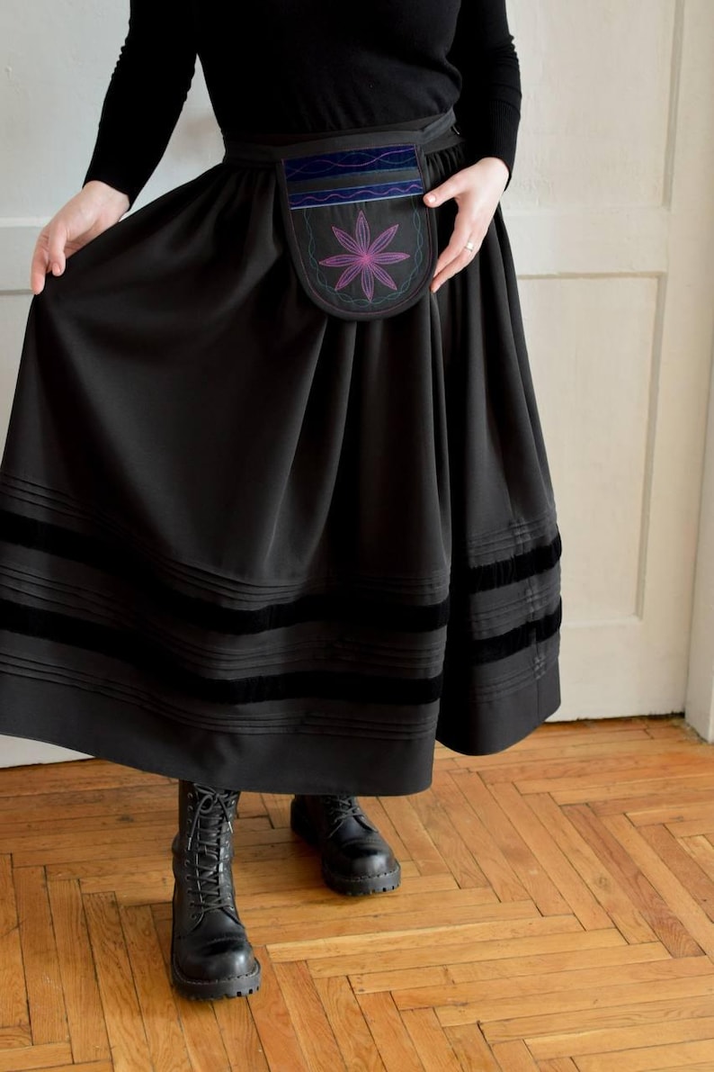 May include: A black midi skirt featuring a pleated waist, two horizontal velvet bands near the hem, and a detachable pouch at the front. The pouch is decorated with a purple and pink embroidered flower motif and blue velvet trim accents.