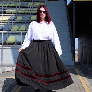 May include: A long black skirt featuring horizontal dark red velvet ribbon trim near the hem, paired with a white long-sleeved top with flared cuffs. The skirt has a gathered waistline and a full, flowing silhouette that reaches the ankles.