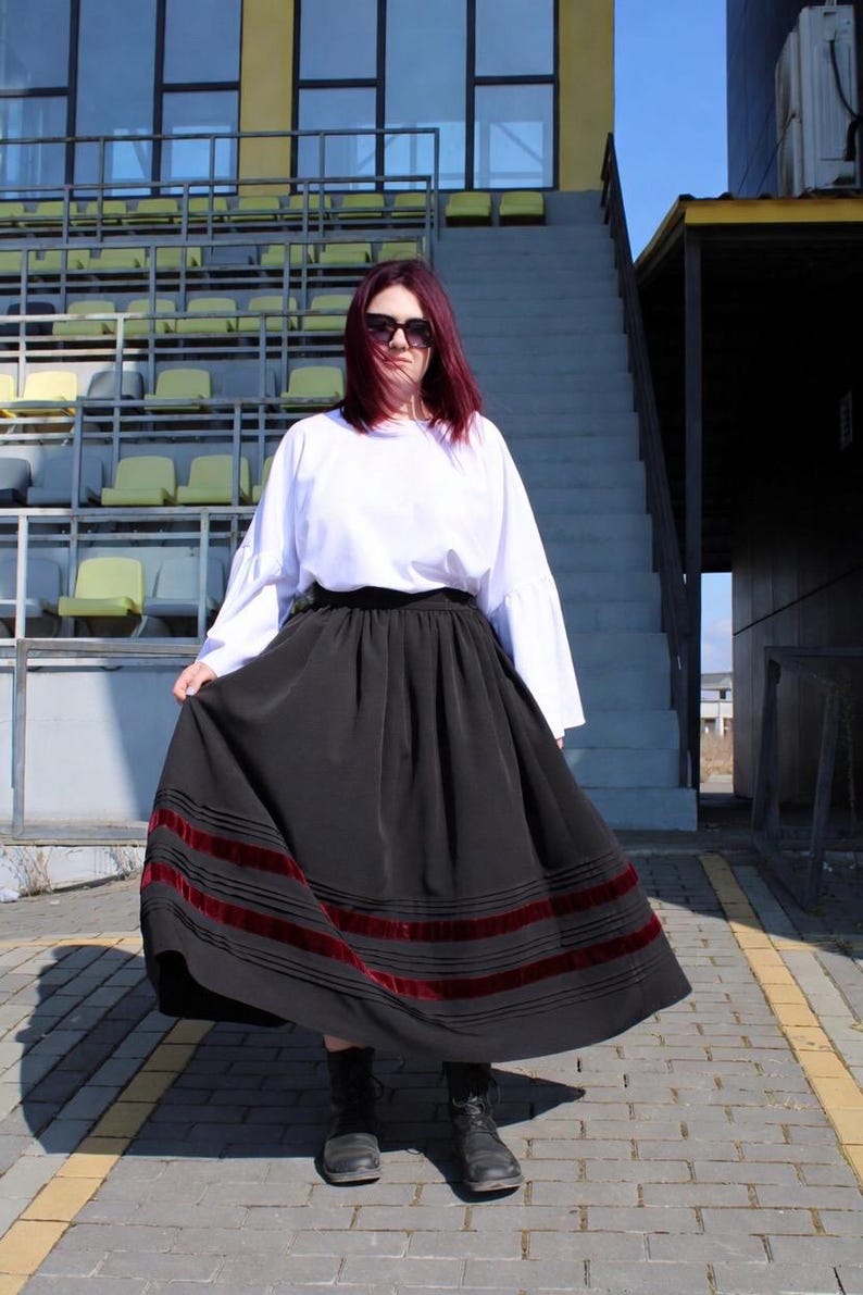 May include: A long black skirt featuring horizontal dark red velvet ribbon trim near the hem, paired with a white long-sleeved top with flared cuffs. The skirt has a gathered waistline and a full, flowing silhouette that reaches the ankles.