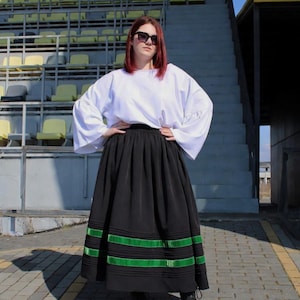 May include: A white long-sleeved blouse with wide sleeves paired with a black ankle-length skirt featuring horizontal green ribbon trim near the hem. The skirt has a gathered waistline and the blouse is tucked in. Black lace-up boots are worn.