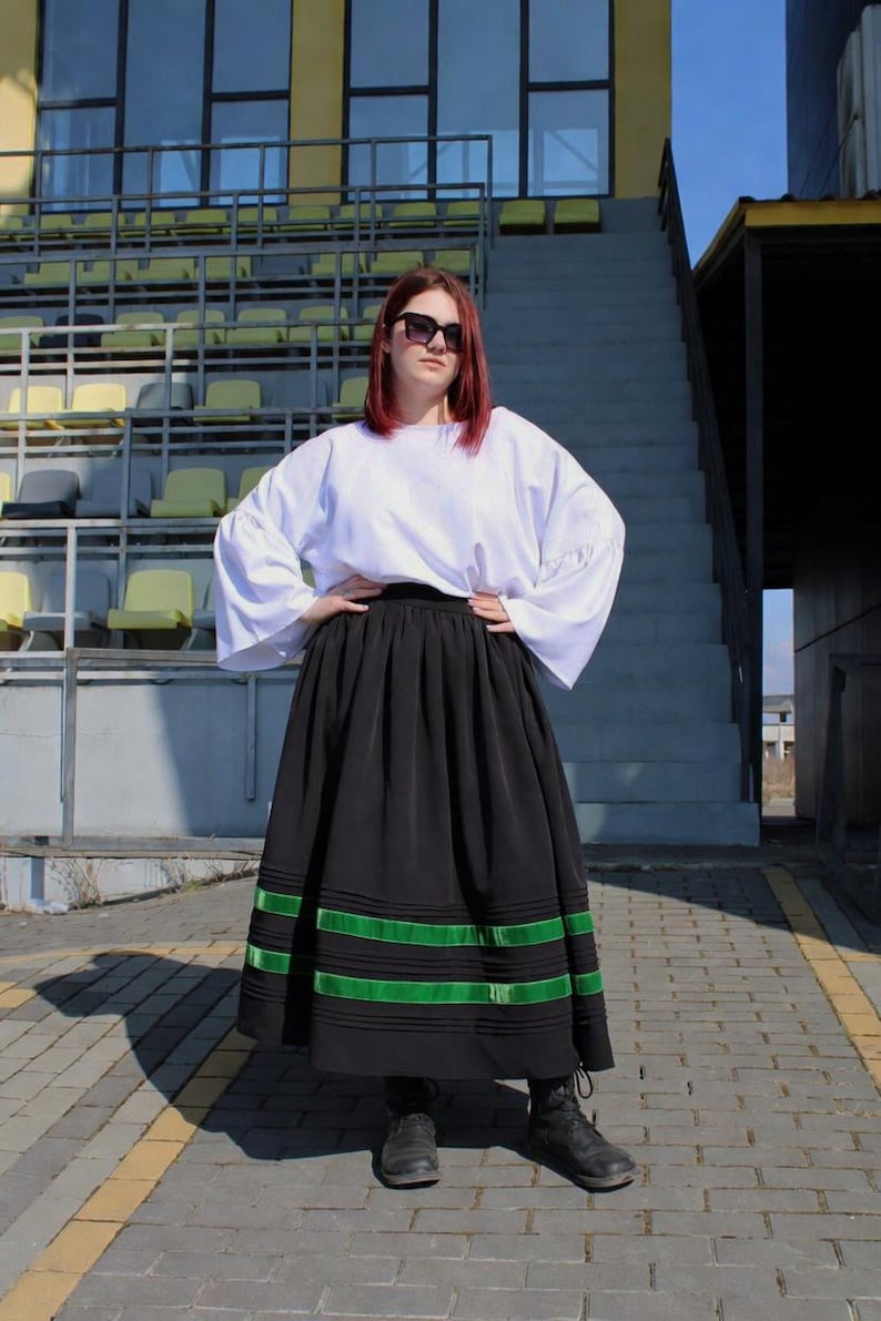 May include: A white long-sleeved blouse with wide sleeves paired with a black ankle-length skirt featuring horizontal green ribbon trim near the hem. The skirt has a gathered waistline and the blouse is tucked in. Black lace-up boots are worn.