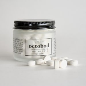 Op de afbeelding: Een glazen pot met een zwarte deksel gevuld met witte tabletten. De pot is gelabeld met "wild mint octobod FLUORIDE-FREE TOOTHPASTE 120 tablets". Witte tabletten liggen verspreid op een wit oppervlak.