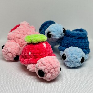 May include: A collection of handmade crochet turtle plushies in various colors. The turtles are primarily pink, red, and blue, with black eyes and a textured, fuzzy appearance. One turtle has a green leaf detail.