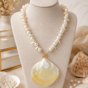 May include: A pearl necklace with a large, pearlescent seashell pendant. The necklace features a strand of white pearls and shell fragments. The pendant is a large, fan-shaped shell with a gradient of yellow and white. The necklace is displayed on a beige bust.