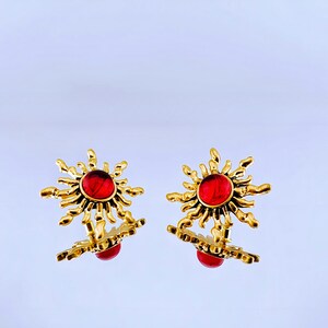 May include: A pair of gold earrings with red gemstone sunburst designs. The earrings are made of metal and have a textured finish.