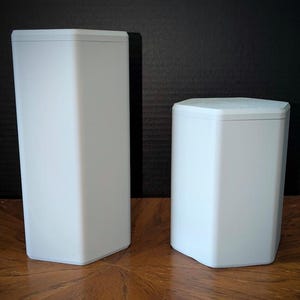 May include: Two white, geometric speakers with a modern design. One speaker is tall and rectangular, while the other is shorter and hexagonal. Both have a clean, minimalist aesthetic, suitable for home audio systems. The speakers are made of a smooth, matte material.