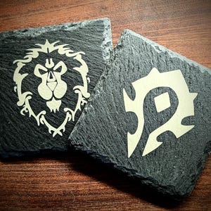 May include: Two square, dark gray slate coasters. One features a lion's head emblem, the other a stylized symbol. Both designs are in a light beige color, creating a contrast against the dark slate. These coasters are ideal for fans of fantasy games.