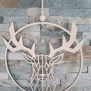 May include: A wooden deer head wall decoration with a geometric design. The deer head is set within a circular frame, with a natural wood finish and black outline. It hangs from a white string and bead. The background is a multi-coloured stone wall.