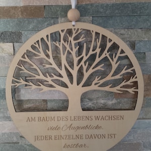 Pode incluir: Decoração de parede redonda em madeira clara com um design de árvore recortada. A árvore tem muitos galhos e é cercada por uma moldura circular. O texto em alemão "AM BAUM DES LEBENS WACHSEN viele Augenblicke. JEDER EINZELNE DAVON IST kostbar." está gravado.