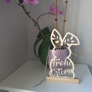 May include: A wooden Easter bunny cutout with the text "Frohe Ostern" on the front. The bunny is decorated with a floral design and is standing on a small wooden base.