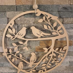 May include: A wooden, circular wall hanging featuring a tree branch design with four birds and leaves. The birds are perched on the branches within the circle. A wooden bead and rope are attached for hanging.