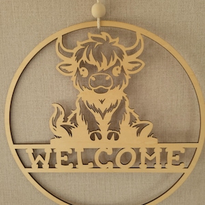 May include: Wooden welcome sign featuring a Highland cow design. The sign is circular with the word "WELCOME" cut out at the bottom. The cow is in the center, with a fluffy coat and horns. A string is attached for hanging.