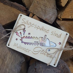 Op de afbeelding: Een rechthoekig houten bord met de tekst "Sägespäne sind Männer Glitzer" en een kettingzaagontwerp. Het bord is vastgebonden met touw en heeft een lichte houten afwerking. De achtergrond is een stapel brandhout.