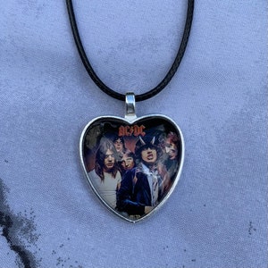 Puede incluir: Un collar con un colgante de corazón plateado y un cordón negro. El colgante presenta una foto de la banda de rock AC/DC.