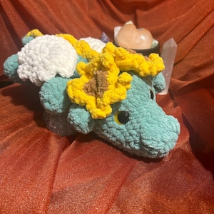 May include: A handmade crochet creature in shades of teal, yellow, and white. The plush toy features a sunflower design on its head and back. The creature has black eyes and is resting on a red surface.