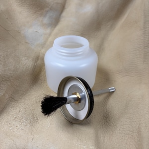 Op de afbeelding: Een doorschijnende witte plastic pot met een wijde opening en een bijpassend schroefdeksel. Het deksel heeft een zwarte rand, een centrale metalen schijf met een messingkleurige moer en een kleine zwarte borstel aan de onderzijde.