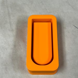 Könnte beinhalten: Eine leuchtend orangefarbene, rechteckige Silikonform mit einer zentralen U-förmigen Vertiefung und abgerundeten Ecken. Die Form weist eine glatte, matte Oberfläche und eine durchgehend gleichmäßige, kräftige orange Farbe auf.