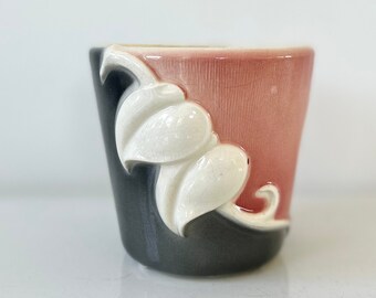 May include: A small, ceramic planter with a pink and gray glaze. The planter has a white, stylized leaf design on the front.