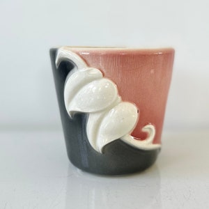 May include: A small, ceramic planter with a pink and gray glaze. The planter has a white, stylized leaf design on the front.