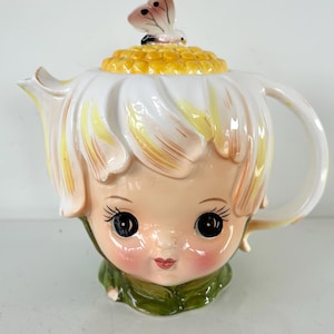 May include: A whimsical ceramic teapot shaped like a flower with a face. The teapot features a white and yellow floral design, a smiling face with large black eyes, and a butterfly on the lid. The handle and spout are integrated into the flower design.