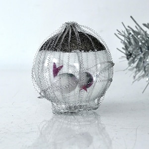 May include: A clear glass Christmas ornament with a silver wire cage and two pink fish inside. The ornament is decorated with glitter.