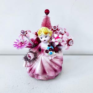 May include: A vintage figurine of a girl in a pink dress, holding a bouquet of flowers. The figurine has blonde hair and is adorned with pink and red floral accents, a small pink tree, and a red ball on top. The base is a light pink color.