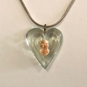 May include: A clear, heart-shaped glass pendant suspended from a silver-toned snake chain. The pendant features a small, beige-colored cameo of a profile portrait embedded in the center, creating a layered effect within the transparent glass.