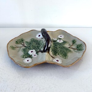 May include: A ceramic serving dish with a light gray base, featuring a central dark brown handle. The dish is decorated with embossed green leaves and white flowers with purple centers. The edges of the dish are scalloped and have a light brown trim.