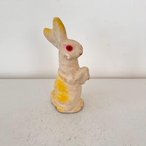 May include: A vintage, off-white rabbit figurine with yellow accents on the lower body and ears. The rabbit has red eyes and is standing upright. The figurine is likely made of plaster or a similar material.
