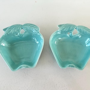 May include: Two turquoise ceramic apple-shaped dishes. Each dish has a leaf design at the top and a small hole. The dishes are glossy and appear to be decorative or for serving small items.
