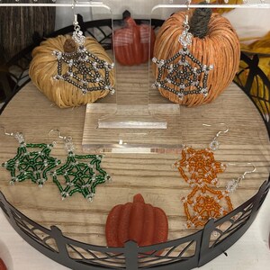 May include: A display of handmade beaded earrings in green, orange, and silver, arranged on a wooden tray with decorative pumpkins. The earrings are spiderweb-like in design, perfect for Halloween or fall. The display includes a black lantern and pumpkin decor.