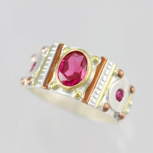May include: A silver, gold, and copper ring with a red gemstone. The ring has a geometric design with a central oval-shaped stone.
