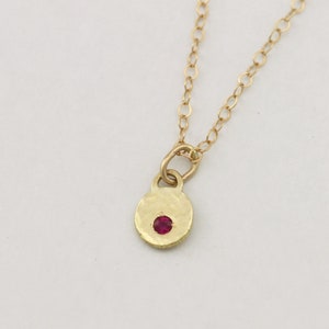 Op de afbeelding: Een gouden ketting met een gehamerde ronde hanger met een kleine rode edelsteen.