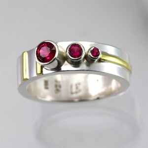 May include: A silver and gold band ring with three red gemstones. The ring has a wide band with a gold stripe.