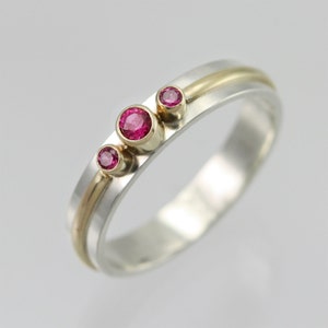 May include: A silver and gold band ring with three pink gemstones. The ring has a thin gold band running along the top.
