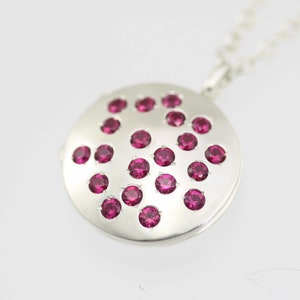 May include: A silver locket necklace with a circular pendant featuring a scattered pattern of small pink gemstones.