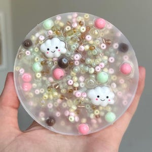 Puede incluir: Un charm redondo de slime transparente con una variedad de cuentas de colores, incluyendo rosa, verde, marrón y amarillo. Hay dos charms de nube blanca con caras sonrientes.