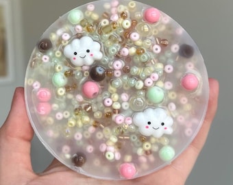 May include: A clear round slime charm with a variety of colorful beads, including pink, green, brown, and yellow. There are two white cloud charms with smiling faces.