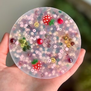 Può includere: Un sottobicchiere rotondo trasparente con una varietà di perline colorate incorporate nella resina. Le perline sono in tonalità di rosa, verde, rosso e marrone. Ci sono anche piccoli ciondoli di plastica di fragole e faccine sorridenti.
