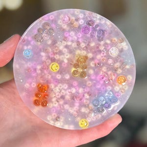 Pode incluir: Um descanso de copo redondo, de resina transparente, com fundo de glitter rosa, branco e iridescente. O descanso de copo é decorado com pequenos amuletos de ursinhos de goma e amuletos de carinhas sorridentes.