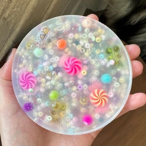 Puede incluir: Un posavasos redondo de resina transparente con cuentas de colores, purpurina y remolinos de caramelo incrustados en el interior.