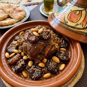 Puede incluir: Un plato de tajine marroquí con un montículo central de carne cocinada a fuego lento, cubierto con ciruelas pasas oscuras, almendras enteras y una pizca de semillas de sésamo. El recipiente de barro color terracota tiene una tapa cónica pintada.