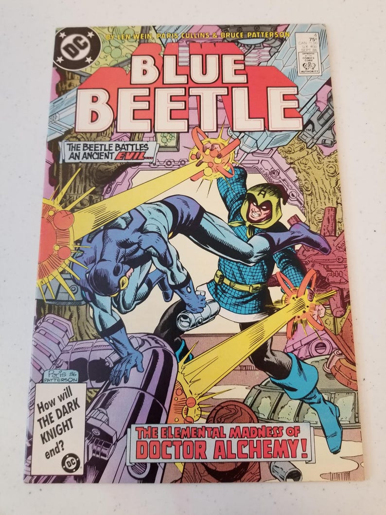 Może przedstawiać: Okładka komiksu z tytułem "BLUE BEETLE" w kolorze czerwonym. Grafika przedstawia dynamiczną scenę walki z Blue Beetle. Okładka zawiera tekst "THE ELEMENTAL MADNESS OF DOCTOR ALCHEMY!" i "How will THE DARK KNIGHT end?"