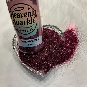 Puede incluir: Un frasco transparente de purpurina color ciruela, etiquetado como "Heavenly Sparkle Glitter Co.", se encuentra sobre un plato en forma de corazón lleno de la misma purpurina. La etiqueta dice "Plum Pixie Dust Fine". La purpurina es de un púrpura profundo y brillante.