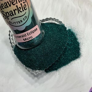 May include: A bottle of Emerald Eclipse Micro glitter from Heavenly Sparkle Glitter Co. is partially tipped over, spilling dark green glitter onto a clear, scalloped dish. The glitter is a vibrant, shimmering color.