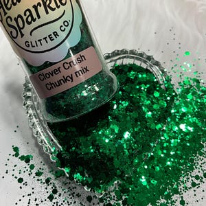 May include: A clear bottle of "Heavenly Sparkle Glitter Co." Clover Crush chunky mix glitter is tilted over a clear, scalloped dish filled with bright green glitter. The glitter is scattered on a white surface.