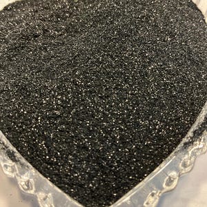 May include: A heart-shaped, clear glass dish filled with black glitter. The glitter sparkles with tiny flecks of silver, creating a shimmering effect. The dish has a scalloped edge.