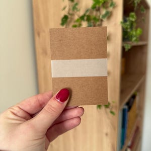Puede incluir: Una tarjeta rectangular marrón con una banda blanca en el medio. La tarjeta se sostiene en una mano con esmalte de uñas rojo. El fondo incluye una estantería de madera y plantas verdes.