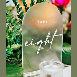 May include: A frosted acrylic table number with a rounded top. The number eight is written in white script on the sign. The sign is sitting on a table with a small white candle in a glass holder.