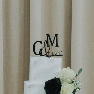 Op de afbeelding: Een witte bruidstaart met drie lagen en een zwarte en gouden taarttopper met de tekst "G&M 11.1.2025". De taart is versierd met een boeket witte en zwarte rozen. Een foto van twee personen staat voor de taart.